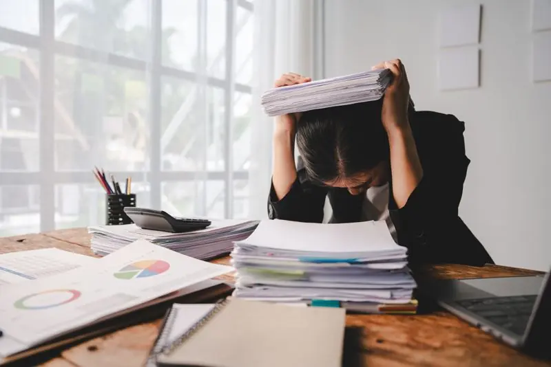Office worker surrounded by printed spreadsheets with a clean business app visible on a screen